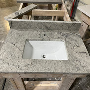 A Stone single vanity top with an under-mount white sink rests on a wooden workbench in a workshop, surrounded by tools and materials.