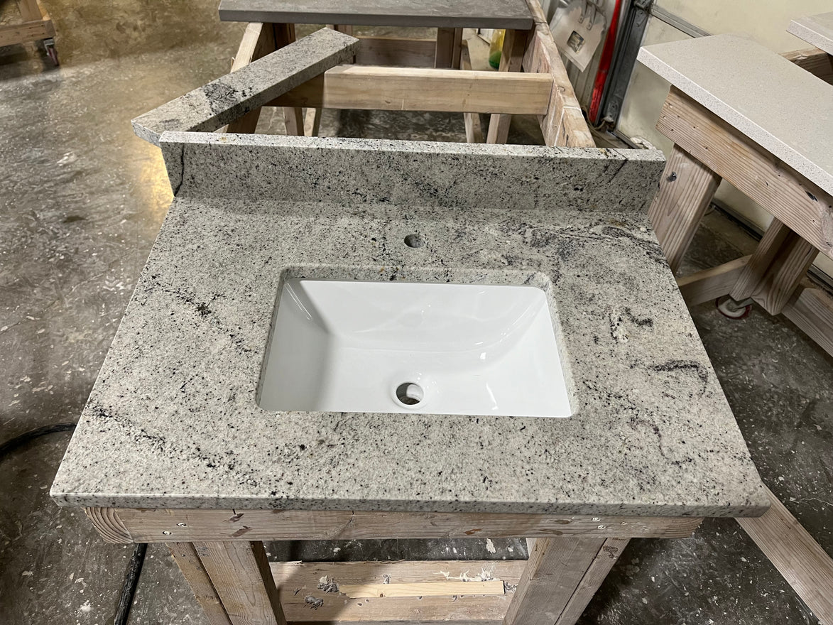 A Stone single vanity top with an under-mount white sink rests on a wooden workbench in a workshop, surrounded by tools and materials.
