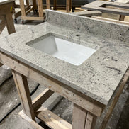 A Stone single vanity top with an integrated white sink rests on a wooden workbench in a workshop, with additional countertops and wooden frames visible in the background.