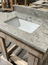 A Stone single vanity top with an integrated white sink rests on a wooden workbench in a workshop, with additional countertops and wooden frames visible in the background.