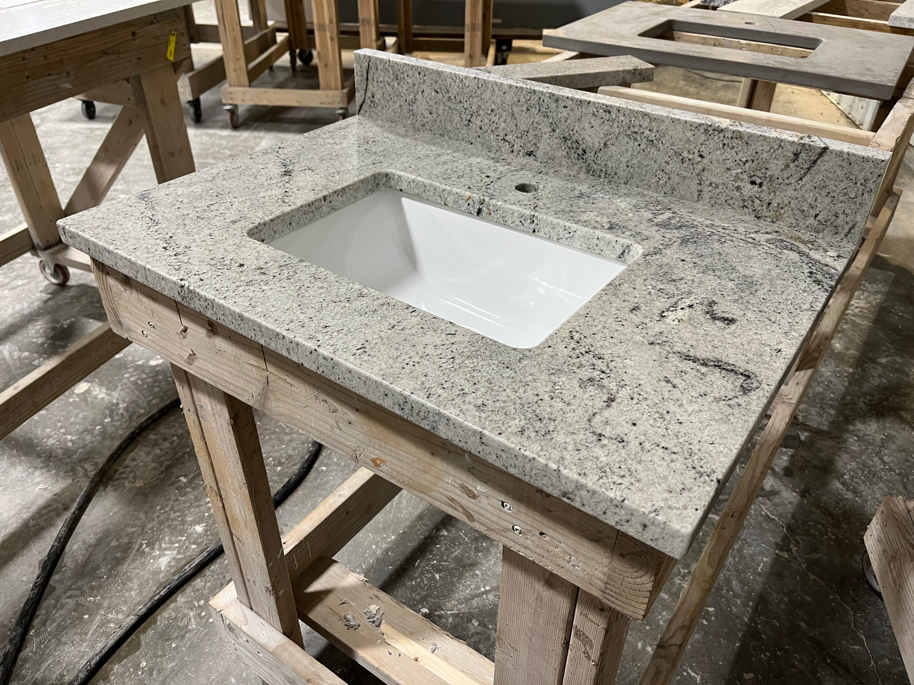 A Stone single vanity top with an integrated white sink rests on a wooden workbench in a workshop, with additional countertops and wooden frames visible in the background.