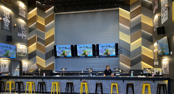 Modern bar interior with yellow and black stools, black bar counter, and multiple televisions on the wall.