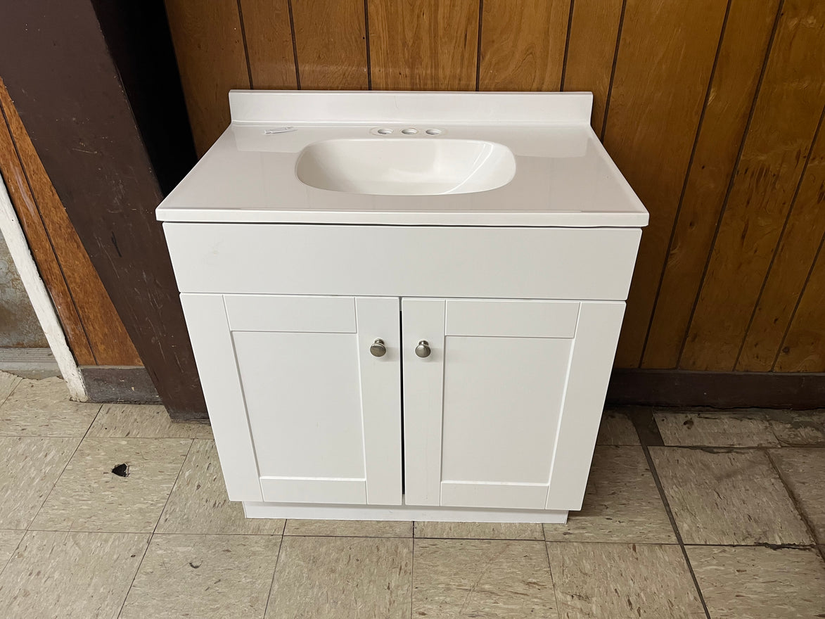 The Vanity with top features a white cabinet with a cultured marble top, built-in sink, and two doors for bathroom storage, shown against a wood-paneled wall on a tiled floor.