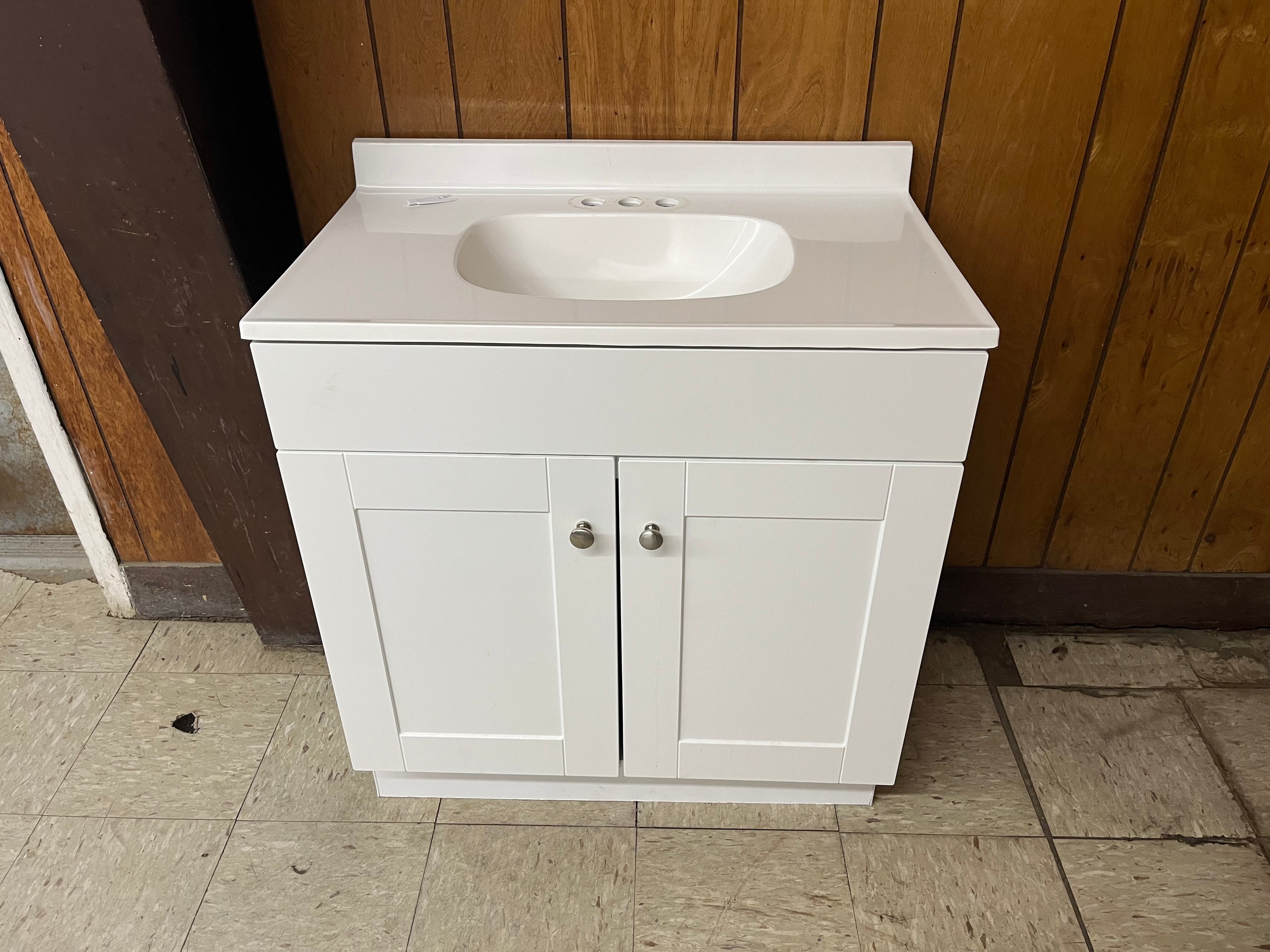 The Vanity with top features a white cabinet with a cultured marble top, built-in sink, and two doors for bathroom storage, shown against a wood-paneled wall on a tiled floor.