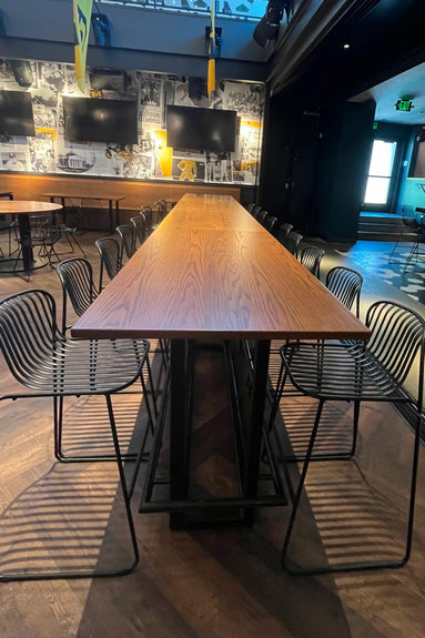 A Custom wood table with black metal chairs is centered in a modern room with TVs and black-and-white sports photos. Sunlight casts chair shadows onto the wooden floor.
