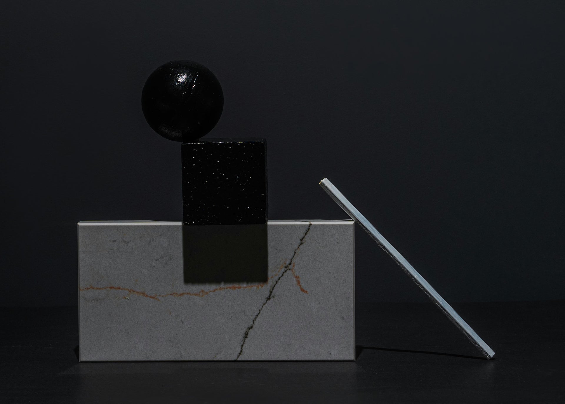 Geometric sculpture with black sphere, cube, and rectangular block on a dark background